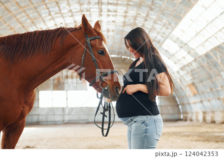 Side view. Beautiful pregnant woman in casual clothes is with the horse on a stable 124042353