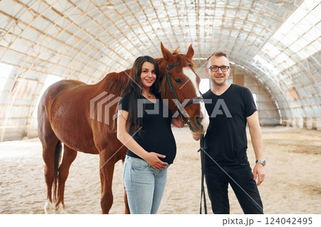 Man with his pregnant wife that is in casual clothes is standing with the horse on a stable Man with his pregnant wife that is in casual clothes is standing with the horse on a stable 124042495