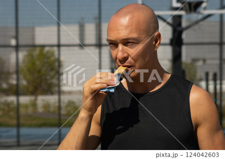 Sports man eating energy bar. Muscular athlete snacks protein bar while resting after workout at stadium. Healthy protein snack, fitness dieting food for sporty lifestyle. 124042603