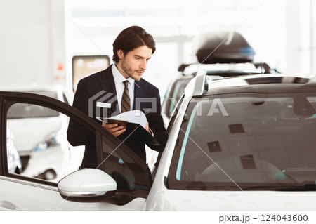 Vehicle Dealership. Car salesperson with clipboard checking auto specifications in local dealership showroom 124043600