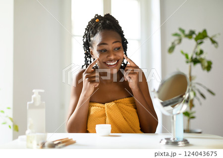 Black young woman in a yellow towel is sitting at a white table, applying skincare to her face. She is looking at her reflection in a mirror and smiling. 124043675