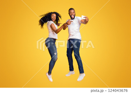 Smiling African American Couple Is Taking Selfie While Jumping, pointing at camera, yellow studio background 124043676