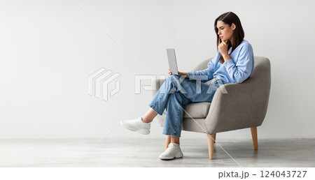 Full length of serious young woman looking at laptop screen, sitting in armchair, having problem with online work or studies against white studio wall, banner with copy space 124043727
