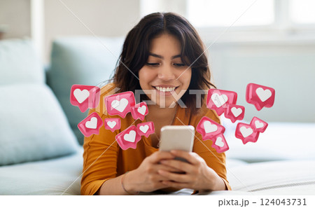 Middle eastern woman lies on a bed, looking down at a smartphone in her hands. She is smiling and appears happy. Above her phone, there are digital heart icons representing likes 124043731