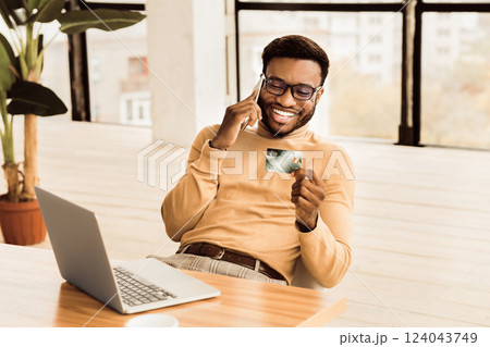 Easy Payments. Happy african worker holding credit card after online purchase and talking on mobile phone. Copy space Easy Payments. Happy african worker holding credit card after online purchase and talking on mobile phone. Copy space 124043749