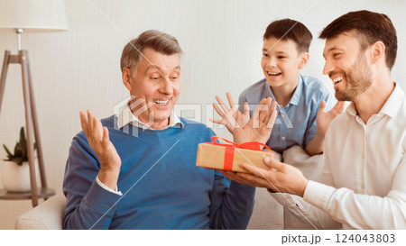 Adult Son And Grandson Giving Grandfather Gift Congratulating Him On His Birthday Sitting On Sofa At Home. Panorama Adult Son And Grandson Giving Grandfather Gift Congratulating Him On His Birthday Sitting On Sofa At Home. Panorama 124043803