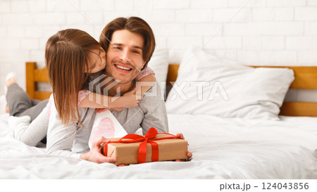 Cute little girl greeting her happy daddy with Father Day, giving him gift box and greeting card, hugging and kissing, laying on bed together at home, free space Cute little girl greeting her happy daddy with Father Day, giving him gift box and greeting card, hugging and kissing, laying on bed together at home, free space 124043856