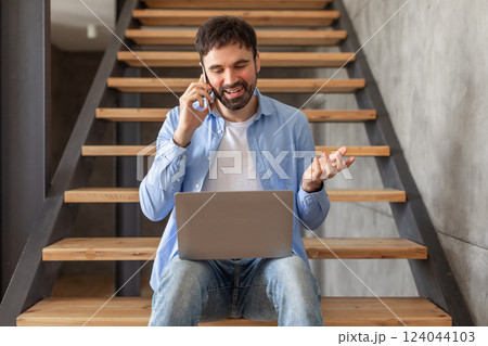 A man is sitting on wooden stairs in a home, talking on a cell phone while holding a laptop on his lap. He is gesticulating with one hand as he speaks, likely discussing something related to his work. A man is sitting on wooden stairs in a home, talking on a cell phone while holding a laptop on his lap. He is gesticulating with one hand as he speaks, likely discussing something related to his work. 124044103