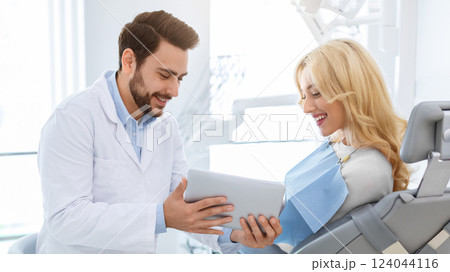 Smiling man dentist showing female patient jaw x-ray on digital tablet, dental clinic interior. Bearded man stomatologist having conversation with lady in dental chair, holding pad, side view Smiling man dentist showing female patient jaw x-ray on digital tablet, dental clinic interior. Bearded man stomatologist having conversation with lady in dental chair, holding pad, side view 124044116