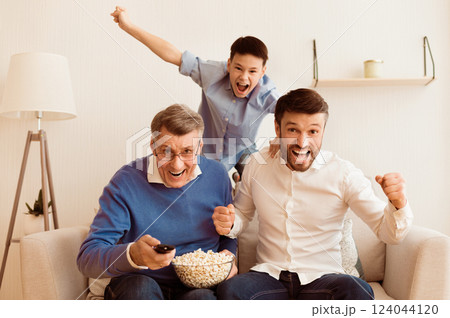 Excited Elderly Man, His Mature Son And Grandson Watching Sports On TV Shouting Celebrating Victory Sitting On Sofa Indoor Excited Elderly Man, His Mature Son And Grandson Watching Sports On TV Shouting Celebrating Victory Sitting On Sofa Indoor 124044120