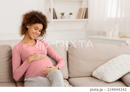 Enjoying pregnancy. Afro girl touching belly and smiling, sitting on couch in the morning, free space 124044142