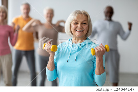 Multiracial group of senior people in sportswear doing strength building fitness exercises with dumbbells, holding fitness tools and smiling at camera, selective focus on positive elderly lady Multiracial group of senior people in sportswear doing strength building fitness exercises with dumbbells, holding fitness tools and smiling at camera, selective focus on positive elderly lady 124044147