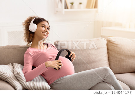 African-american pregnant woman holding headphones to belly and listening to music in lving room, free space 124044177
