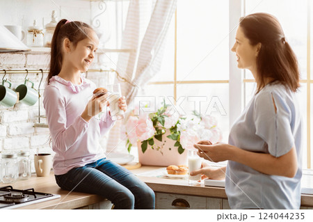 A young girl, wearing a pink long-sleeved shirt and jeans, sits on a kitchen counter, holding a muffin and a glass of milk. She is looking to her right, where is her mother standing A young girl, wearing a pink long-sleeved shirt and jeans, sits on a kitchen counter, holding a muffin and a glass of milk. She is looking to her right, where is her mother standing 124044235