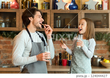 Snack Time. Cheerful dad and little daughter eating fresh baked cookies and drinking milk in kitchen, enjoying homemade pastry, wearing aprons and smiling to each other, having fun at home, copy space Snack Time. Cheerful dad and little daughter eating fresh baked cookies and drinking milk in kitchen, enjoying homemade pastry, wearing aprons and smiling to each other, having fun at home, copy space 124044427