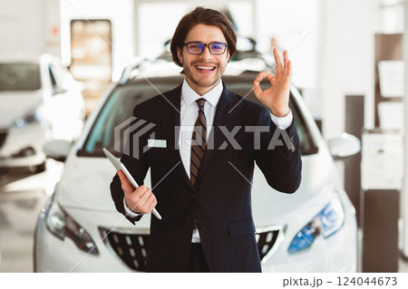 Car Sales. Smiling Handsome Salesman In Glasses Gesturing Okay Standing In Luxury Auto Store. Free Space 124044673