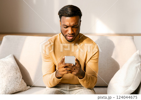 Discontended African American Man Holding Smaprtphone And Frowning Reading Message Sitting On Sofa Indoor. Selective Focus 124044675
