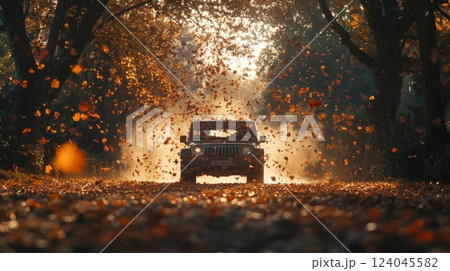 A jeep moves along a dusty road during autumn, surrounded by trees with vibrant leaves. The air is filled with swirling autumn foliage as sunlight filters through the branches. 124045582