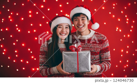 A young couple wearing Santa hats smile at the camera, holding a gift box wrapped in silver paper. 124046730