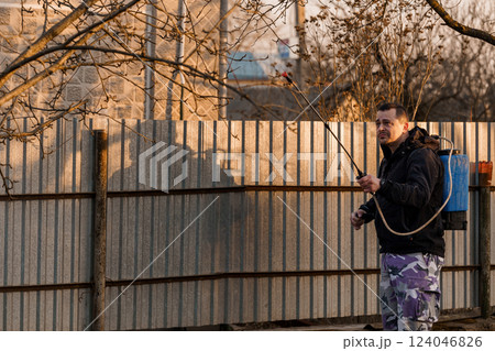 Man in Purple Camouflage Pants Spraying Chemicals in Backyard During Evening Sunset with Silhouetted Fence and Leafless Trees Man in Purple Camouflage Pants Spraying Chemicals in Backyard During Evening Sunset with Silhouetted Fence and Leafless Trees 124046826
