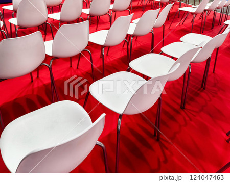Rows of empty white chairs arranged on a vibrant red carpet for an upcoming event at a convention center Rows of empty white chairs arranged on a vibrant red carpet for an upcoming event at a convention center 124047463