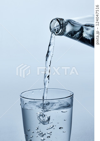 Close-up photo of water being poured from glass bottle into clear glass, showing flowing stream and splash against light blue background. 124047516