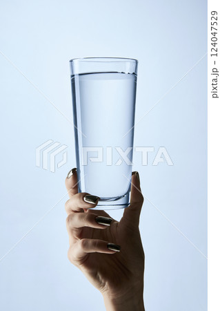 Close-up photo of hand with manicured nails holding clear glass filled with water, against soft blue background. Mock up. 124047529