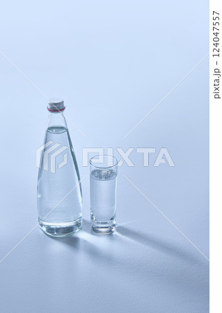 Clear glass bottle and matching glass, both filled with transparent water, on light blue surface, casting long shadows. Mock up. 124047557