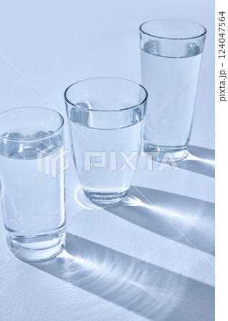 Three glasses filled with water stand on light surface, casting long, blurry shadows. Light refracts through water, creating sparkling effect. Mock up. Three glasses filled with water stand on light surface, casting long, blurry shadows. Light refracts through water, creating sparkling effect. Mock up. 124047564