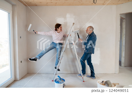 Senior husband and wife standing on a ladder repainting the walls. 124049051