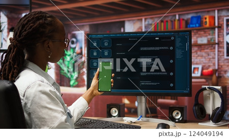 Woman using isolated screen smartphone and AI LLM chatbot on computer screen in home office. Person holding chroma key phone uses artificial intelligence chatbot on monitor, camera A 124049194
