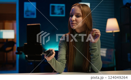 Young girl recording introductory video with cellphone on selfie stick, inviting viewers to subscribe to channel. Media star in home studio uses mobile phone to film herself, camera B 124049264