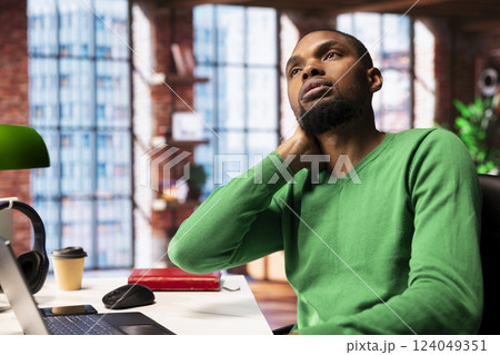 Remote worker in home office stretching neck after sitting at desk too long. Teleworker in living room doing muscle exercises after feeling pain from uncomfortable posture Remote worker in home office stretching neck after sitting at desk too long. Teleworker in living room doing muscle exercises after feeling pain from uncomfortable posture 124049351
