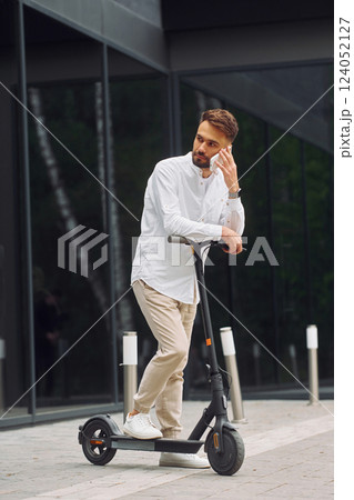 Talking by phone. Stylish man is with his scooter outdoors at daytime Talking by phone. Stylish man is with his scooter outdoors at daytime 124052127