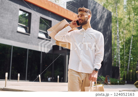 Holding bags and talking by phone. Stylish young man have shopping day. Outdoors with bags 124052163