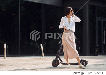 Using phone. Beautiful young woman is with her scooter outdoors 124052211
