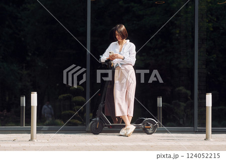Using phone. Beautiful young woman is with her scooter outdoors 124052215