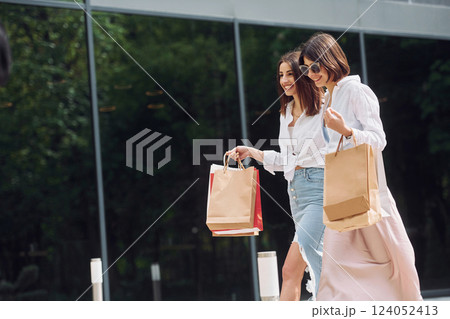 Near modern building. Two female friends have a shopping day. Walking outdoors with bags 124052413