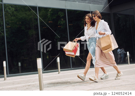Near modern building. Two female friends have a shopping day. Walking outdoors with bags 124052414