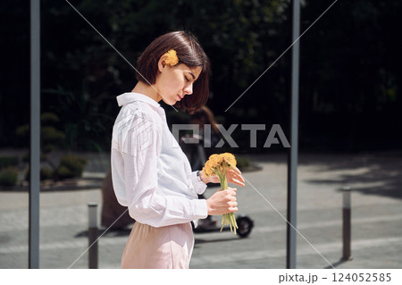Against building with big windows. Beautiful brunette is standing outdoors with flowers in her hands Against building with big windows. Beautiful brunette is standing outdoors with flowers in her hands 124052585
