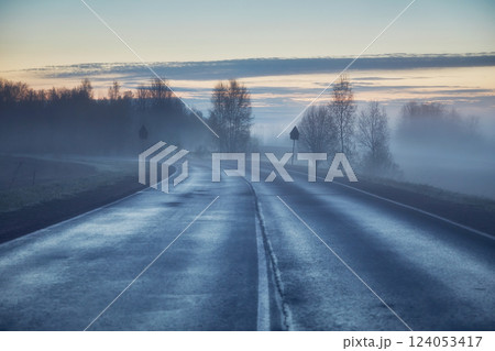 Asphalt road in the countryside early in the morning at blue hour. In the background there are trees, sky and fog. 124053417