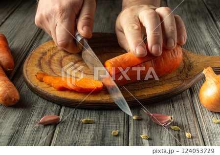 A person skillfully cuts bright orange carrots on a rustic wooden board, surrounded by garlic and cardamom pods, depicting a cozy kitchen atmosphere A person skillfully cuts bright orange carrots on a rustic wooden board, surrounded by garlic and cardamom pods, depicting a cozy kitchen atmosphere 124053729