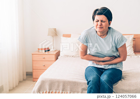 Mature caucasian female experiencing stomach pain while sitting on bed in bedroom 124057066