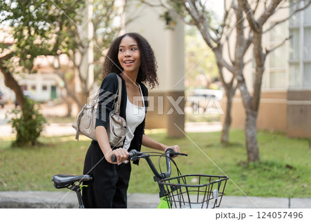 Joyful Exploration and Eco-Friendly Transport. A young woman smiles while walking with her bicycle through a sunny urban landscape. 124057496