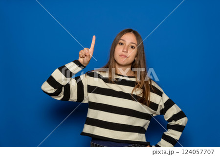 Hispanic teen girl about 15 years wearing striped sweater over blue background pointing finger up and looking inspired by a brilliant thought, showing good idea sign, having a smart solution in mind. 124057707