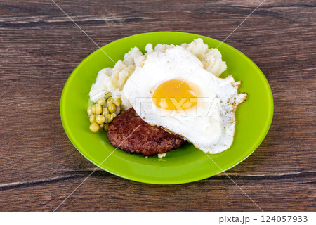 Green plate with a cutlet under a fried egg with mashed potatoes and green peas. 124057933