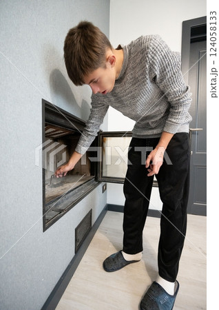 Young man tending to a modern fireplace in a contemporary living room with light gray walls and minimalistic decor while dressed in casual attire during a quiet afternoon Young man tending to a modern fireplace in a contemporary living room with light gray walls and minimalistic decor while dressed in casual attire during a quiet afternoon 124058133