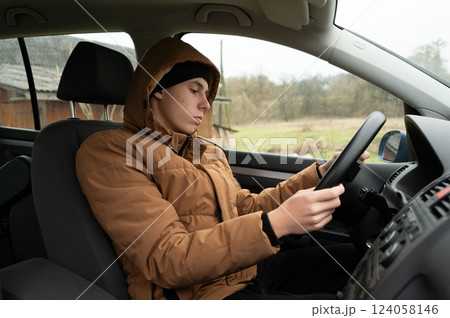 Young driver focused on the road inside a cozy car, surrounded by a calm rural landscape on a grey, cloudy day 124058146