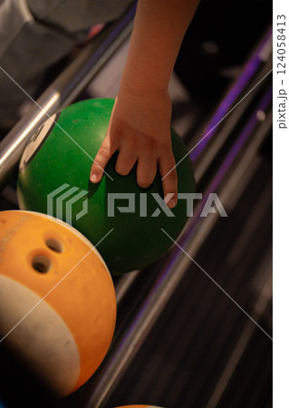 Hand reaching for vibrant bowling ball in colorful, well-lit bowling alley with various balls and blurred background creating dynamic atmosphere 124058413