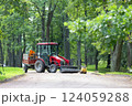 A red tractor with a trailer is parked on a gravel path in a park. Its bucket is filled with gravel. Repairing paths in a park. Preparing for the tourist season. 124059288